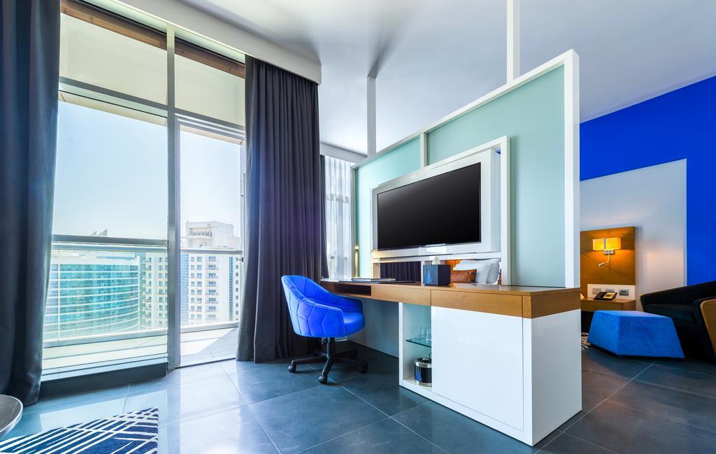 Tryp By Wyndham Dubai Hotel Exterior foto The photo shows a modern room with large windows allowing natural light to enter. There's a stylish desk area featuring a flat-screen TV mounted on the wall, along with a light wooden desk and a blue chair. The wall behind the TV is painted in a ligh